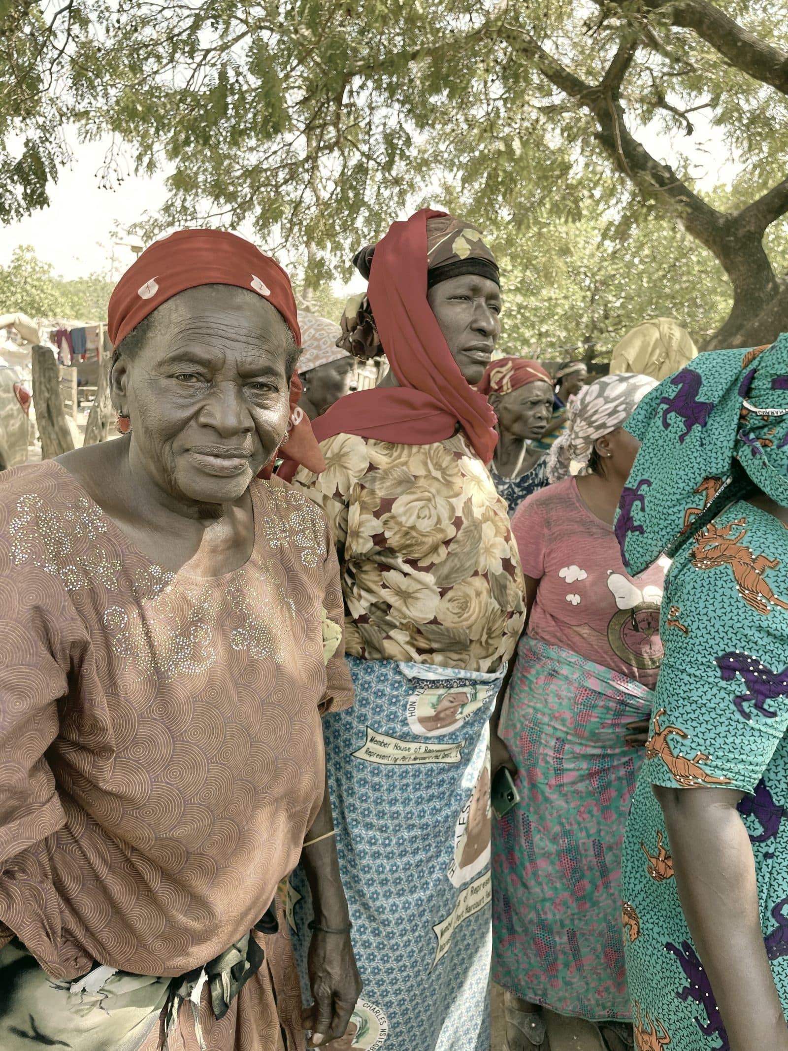 Women of an Abuja IDP camp community gathered beneath a tree, dressed in vivid patterned fabric