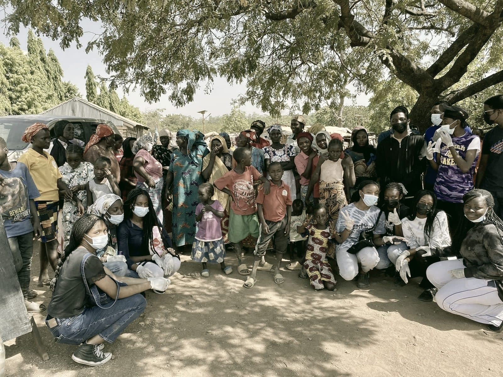 Wide view of families and volunteers standing beneath the tree canopy at the camp