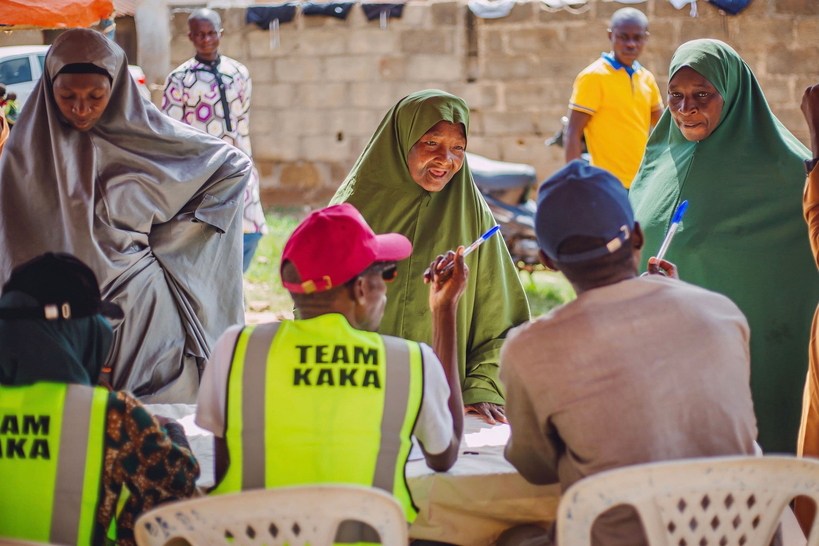 Women speaking across a table while members of a seated team write during an outdoor community meeting