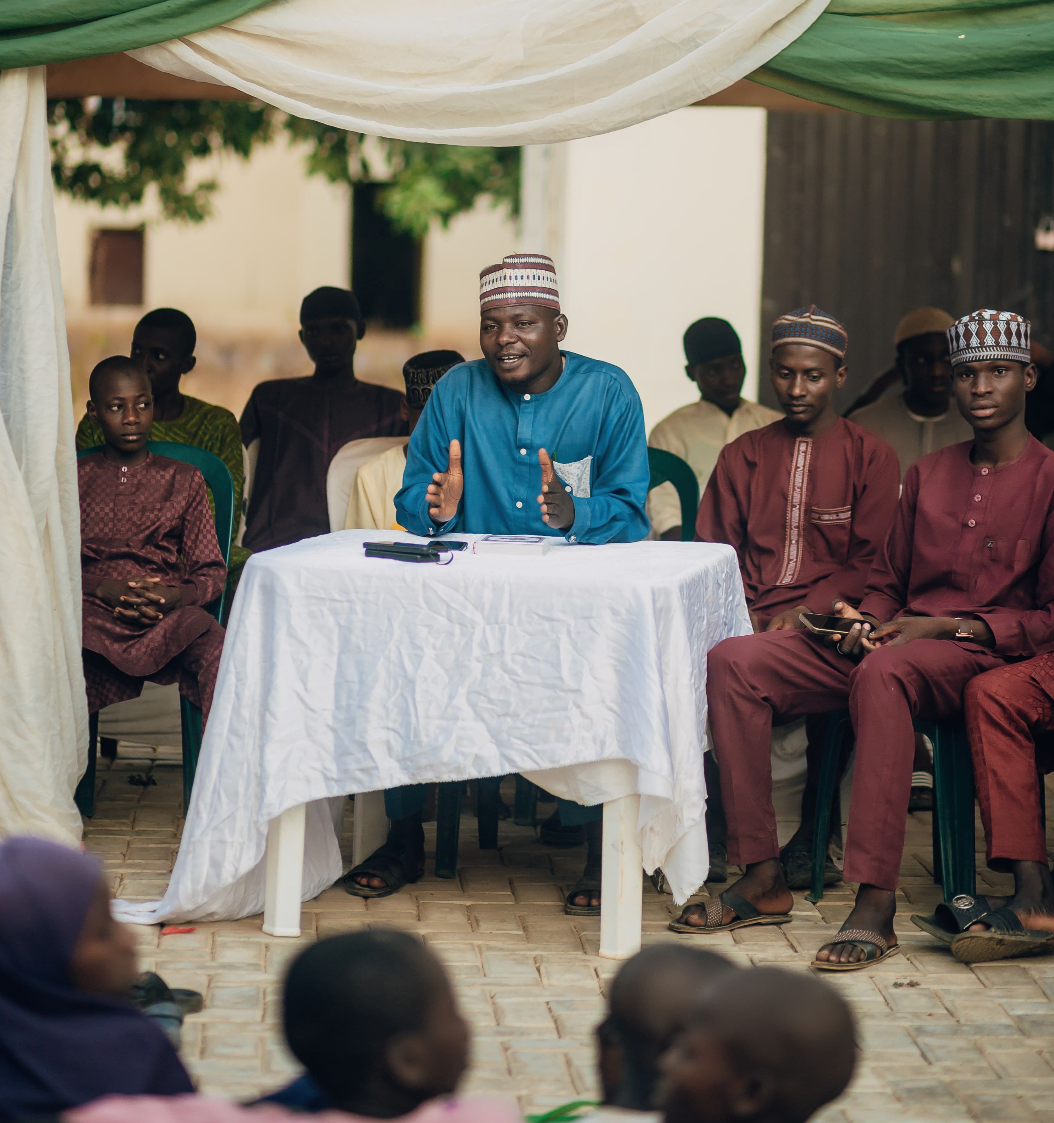 A man in blue speaking at a table while a seated outdoor gathering listens beneath a canopy