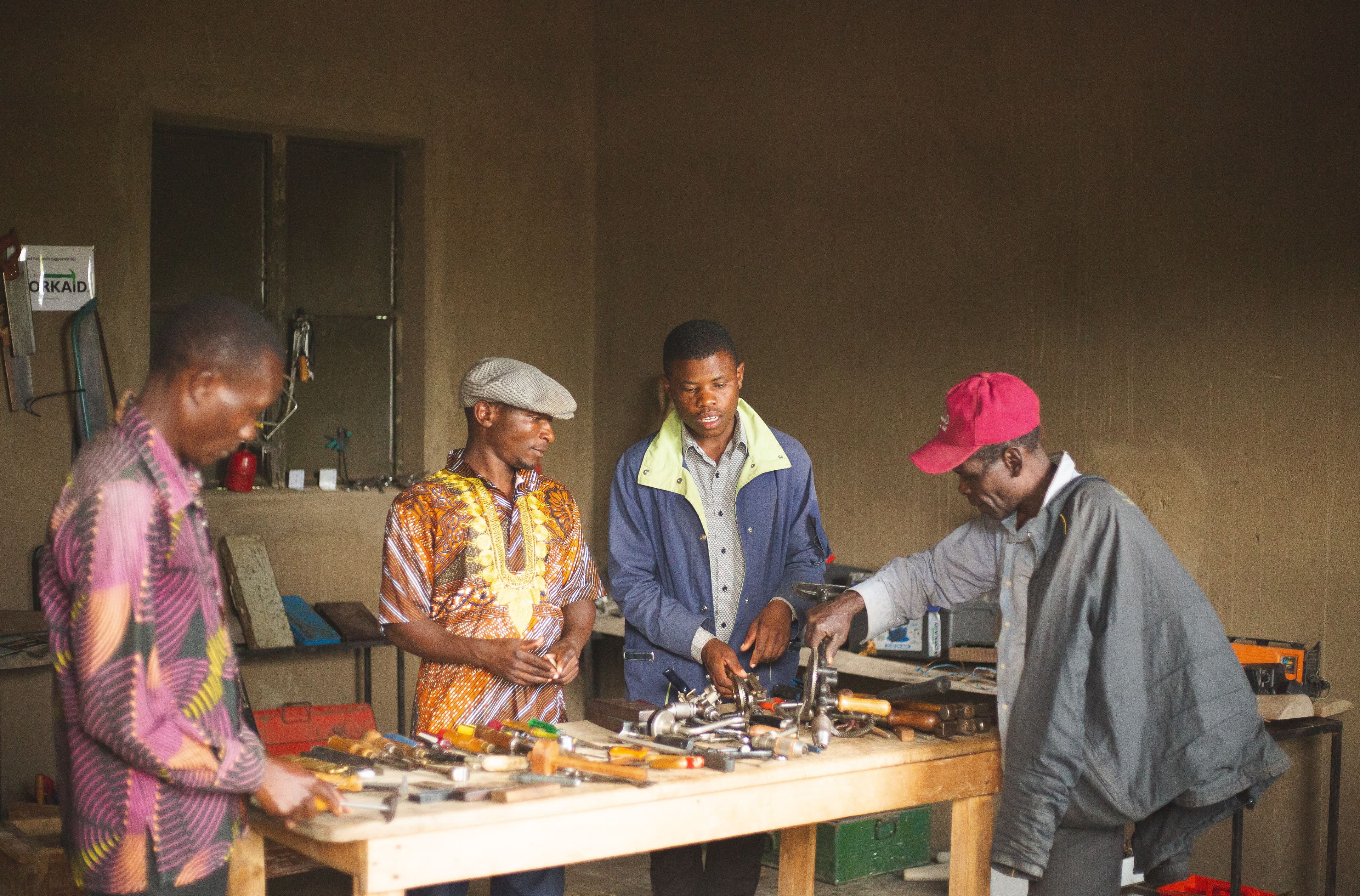 Four men gathered around hand tools and equipment at a workshop bench